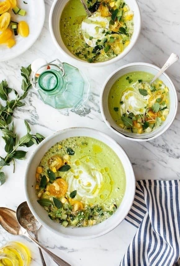 Three bowls of green soup topped with herbs, yellow cherry tomatoes, and a dollop of cream, garnished with fresh herbs and accompanied by a striped napkin