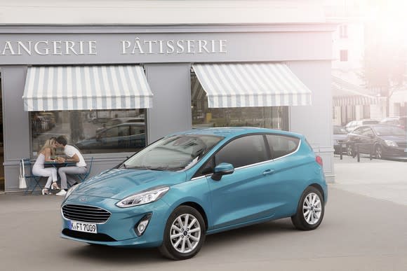 A teal two-door Ford Fiesta hatchback is parked in front of a French bakery.