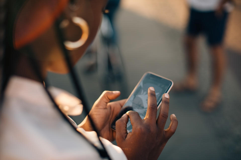 Person uses a smartphone outdoors, tapping the screen with fingers. Unidentifiable because the face is not shown. Multiple people are blurred in the background