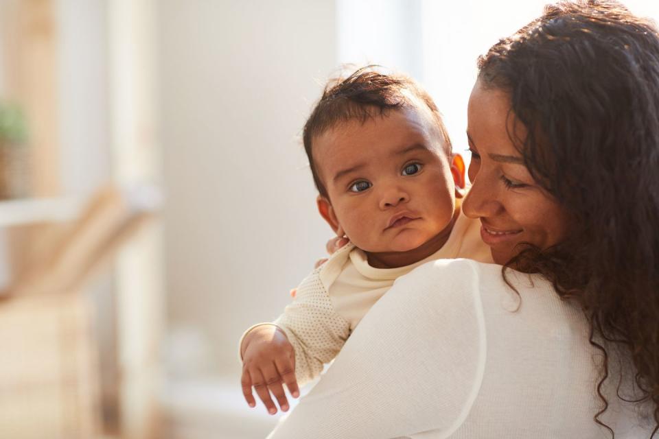 smiling loving young mom with curly hair standing in room and holding adorable baby boy with chubby cheeks