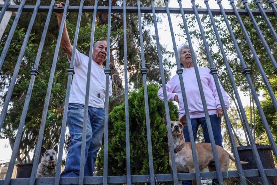 An elderly couple look out through the iron gate of their rural property.