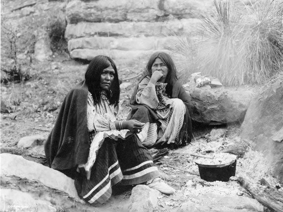 Two Apache Indian women sit in front of a campfire, a cooking pot in front of one.