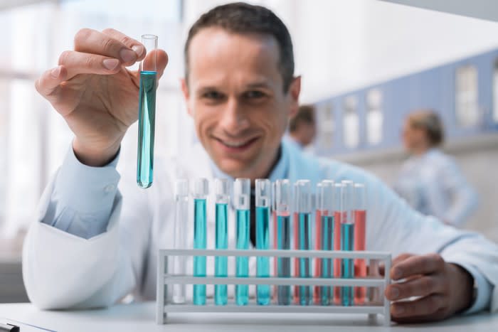 A man in a lab coat smiling and holding test tube with a rack of test tubes in front of him.