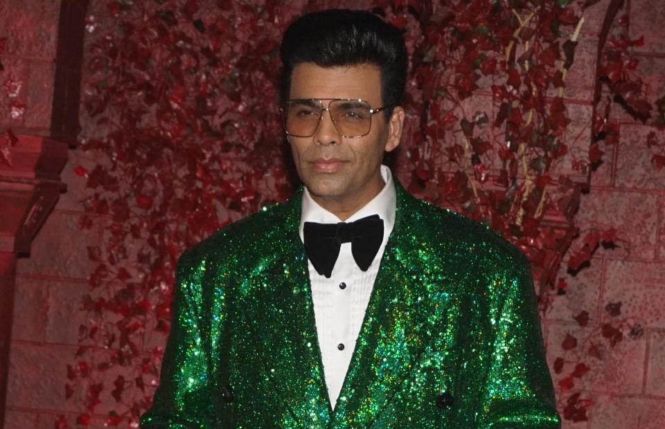 Indian filmmaker Karan Johar in a sparkly emerald green suit, black bowtie, and glasses, standing against a red brick backdrop with bright red leaves decorating it.