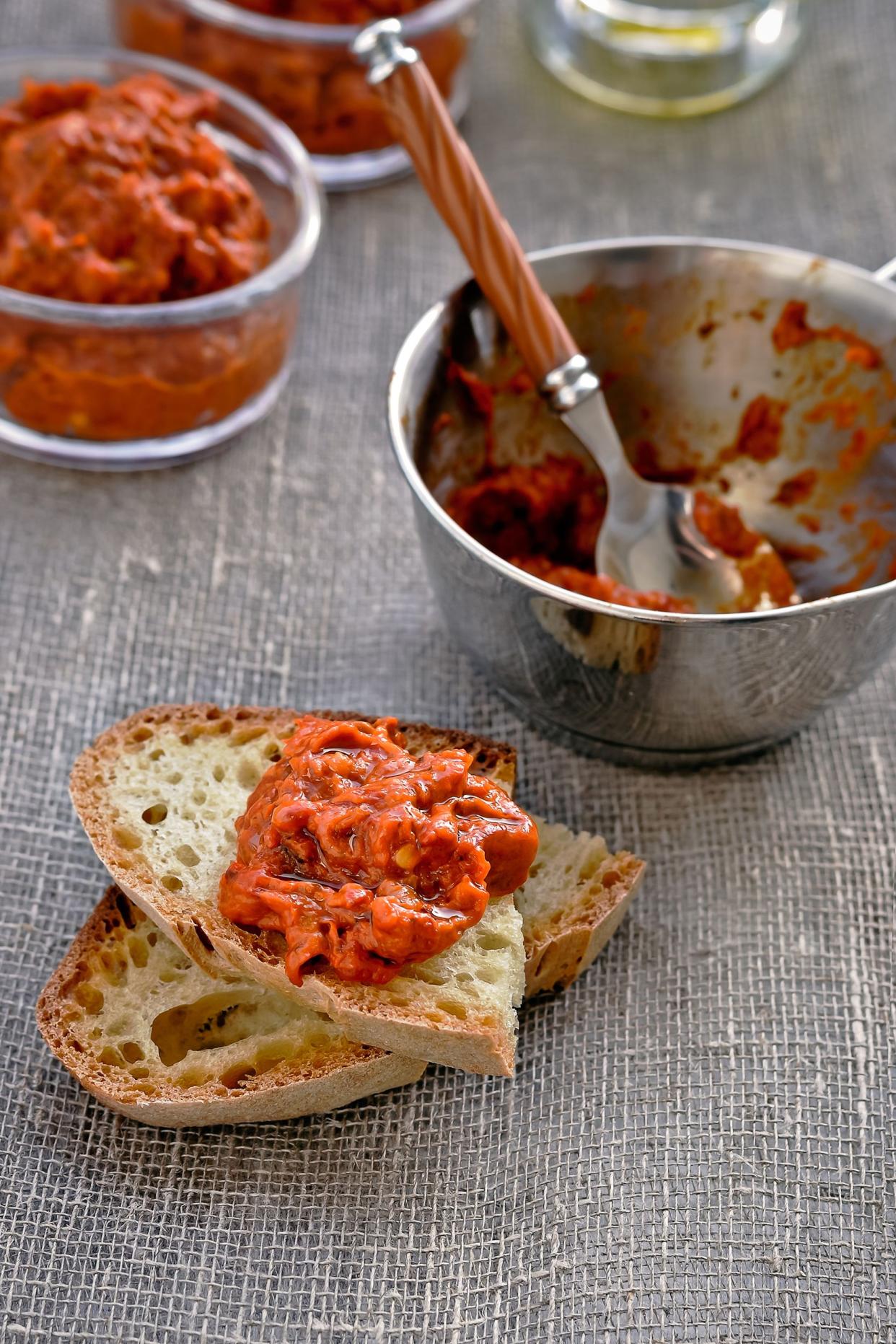 Red pepper relish on a piece of bread with relish in a stainless steel bowl with a spoon with relish in two glass round bowls