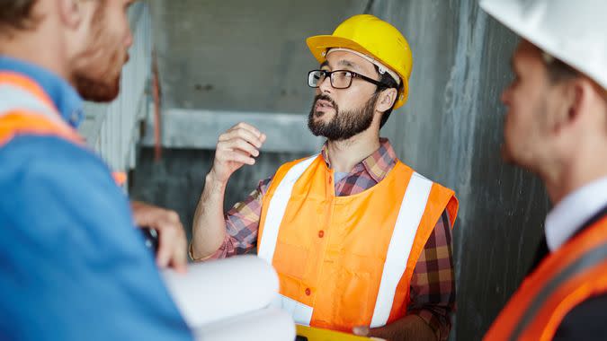 Portrait of bearded expert explaining construction details to workmen, making measurements as they go, all wearing reflective orange vests and hard hats.