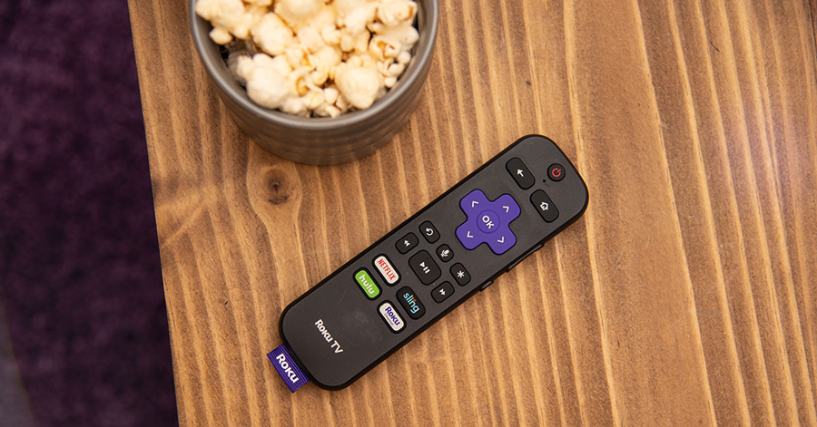 A Roku TV remote on a table next to a bowl of popcorn.