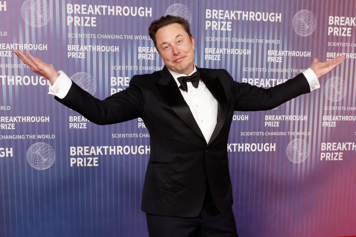 Elon Musk wearing a suit and bowtie whilst shrugging with his hands in the air