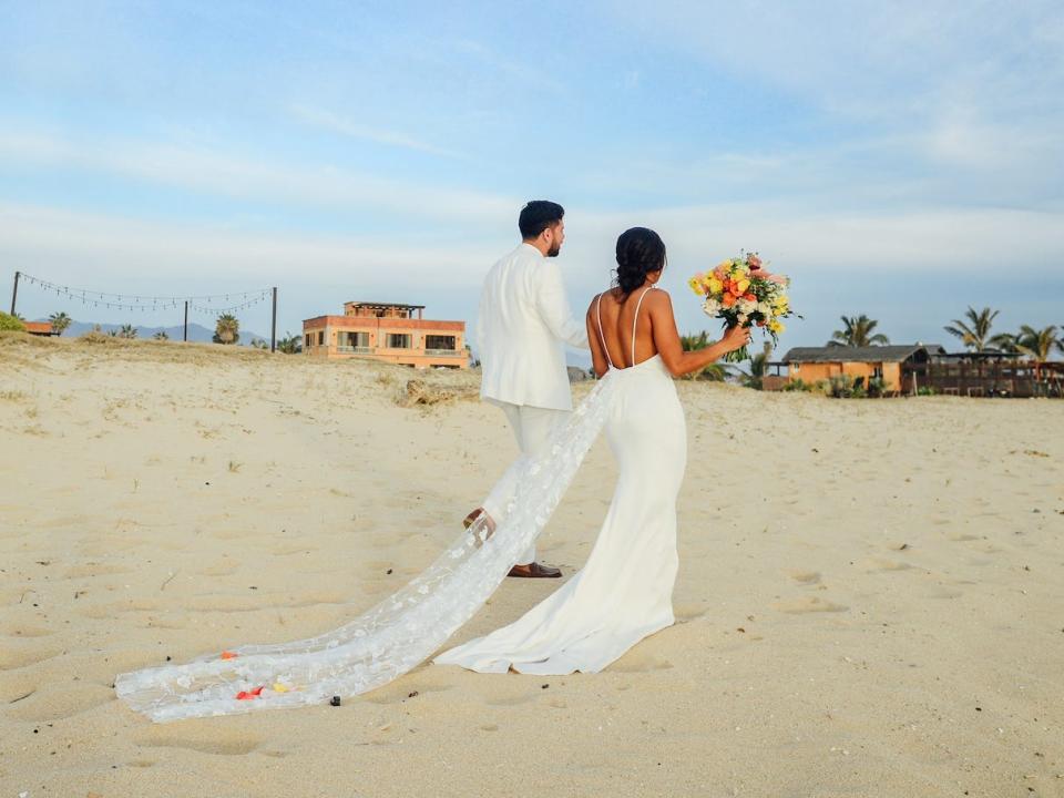 A man and woman walk on a beach in their wedding attire.