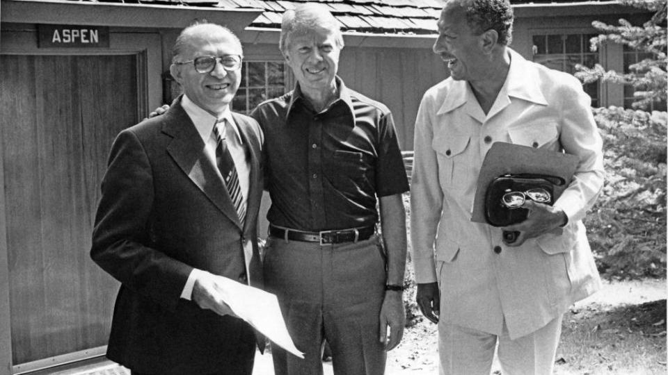 israeli prime minister menachem begin, us president jimmy carter, and egyptian president anwar al sadat stands outside a wooden building and smile, they wear business attire