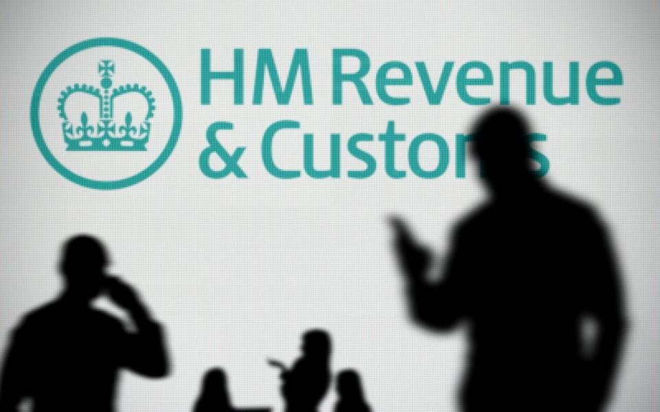 HMRC logo is seen on an LED screen in the background while a silhouetted person uses a smartphone in the foreground