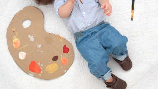 baby in bob ross 80s halloween costume with diy cardboard palette, wig 'fro, paint brushes, jeans, casual button up shirt