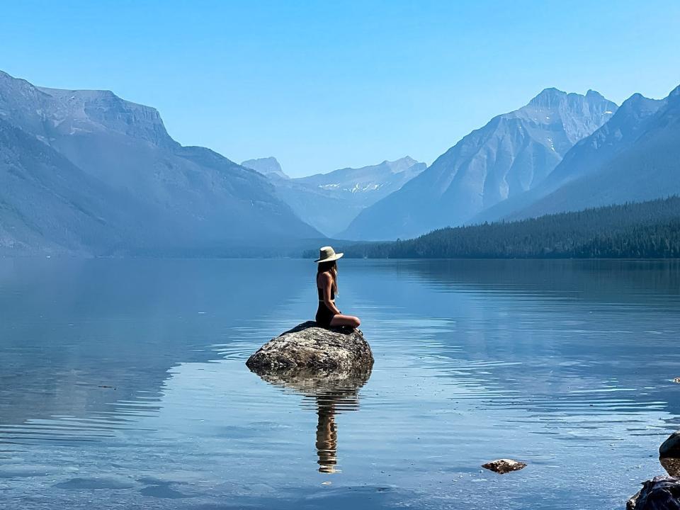 Emily sitting on a rock in the middle of a large body of water surrounded by water in Glacier National Park.
