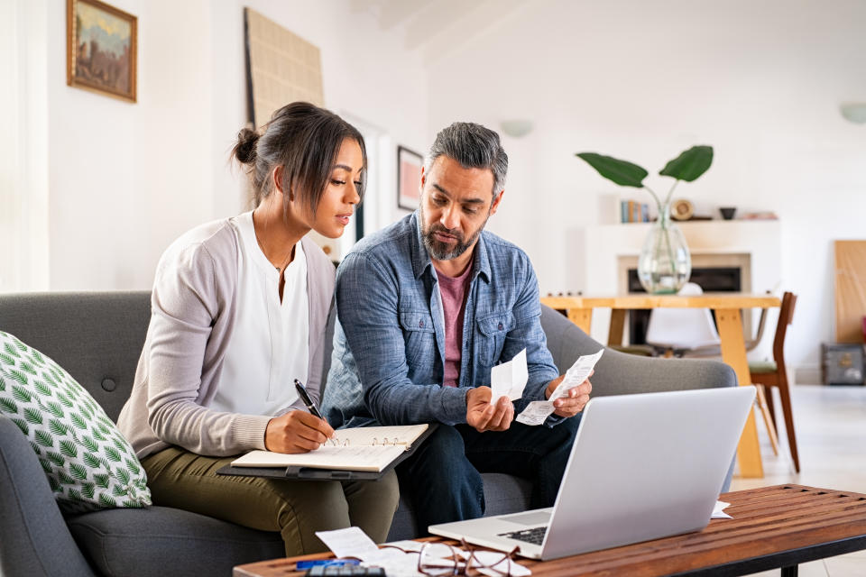 Mature couple looking at how to save money at home using laptop and calculator. Multiethnic couple working on computer while calculating finances sitting on couch. Mature indian man with african american woman at home analyzing their finance with documents.