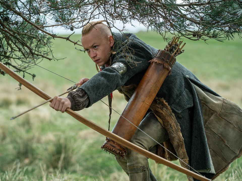 Ian Murray drawing a bow with an arrow notched in it while crouching in the grass with a quiver of arrows slung over his shoulder.