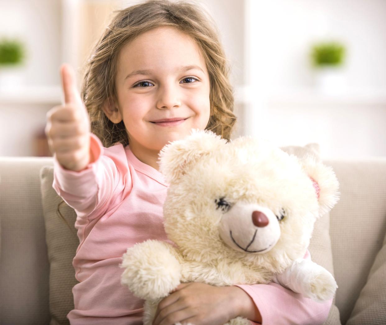 little girl sitting on a sofa with teddy bear and showing thumbs up
