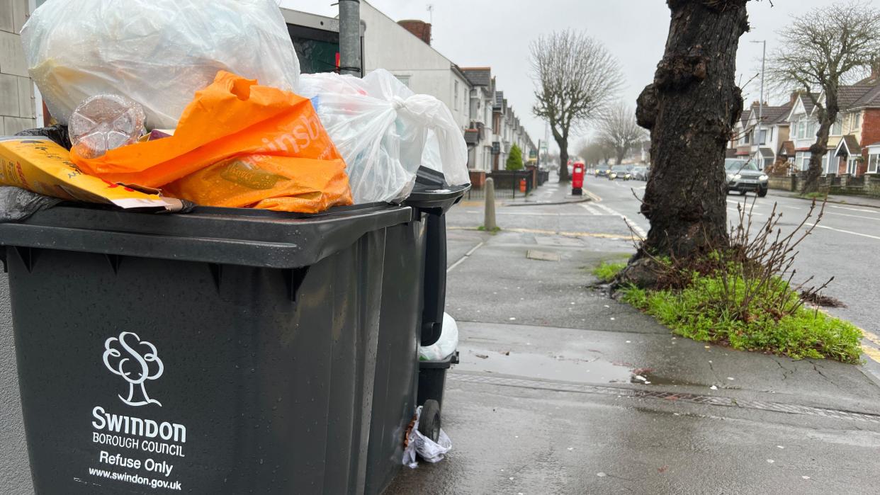 Overflowing black wheelie bins on a pavement on a rainy day with a Swindon Borough Council logo