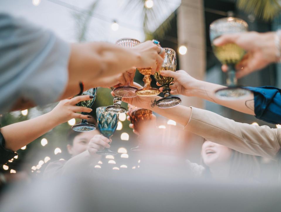 Group of people raise decorative glasses in a celebratory toast outdoors, blurred lights and palm trees in the background