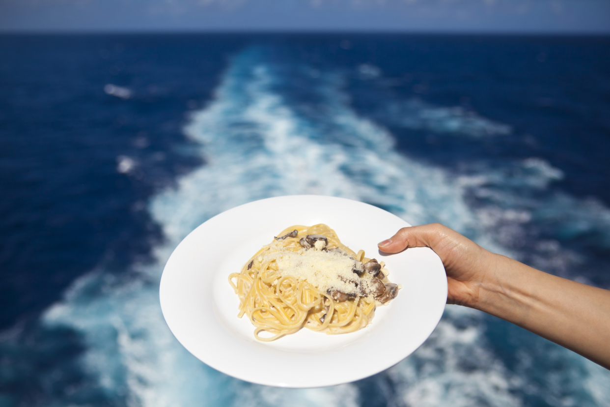 Female right hand holding a dish of spaghetti, selective focus, against the trace of the ship on the ocean on a cruise ship