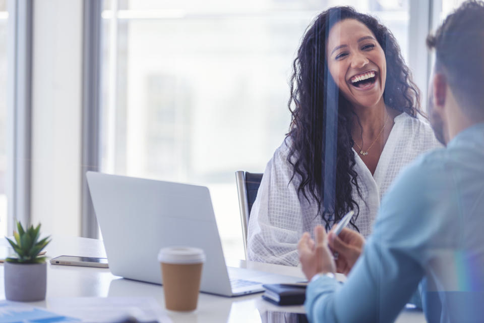 Business colleagues having a conversation. They are both young business people casually dressed in a bright office. Could be an interview or consultant working with a client. She is laughing and having fun. There is a laptop computer on the table  One person has his back to us.