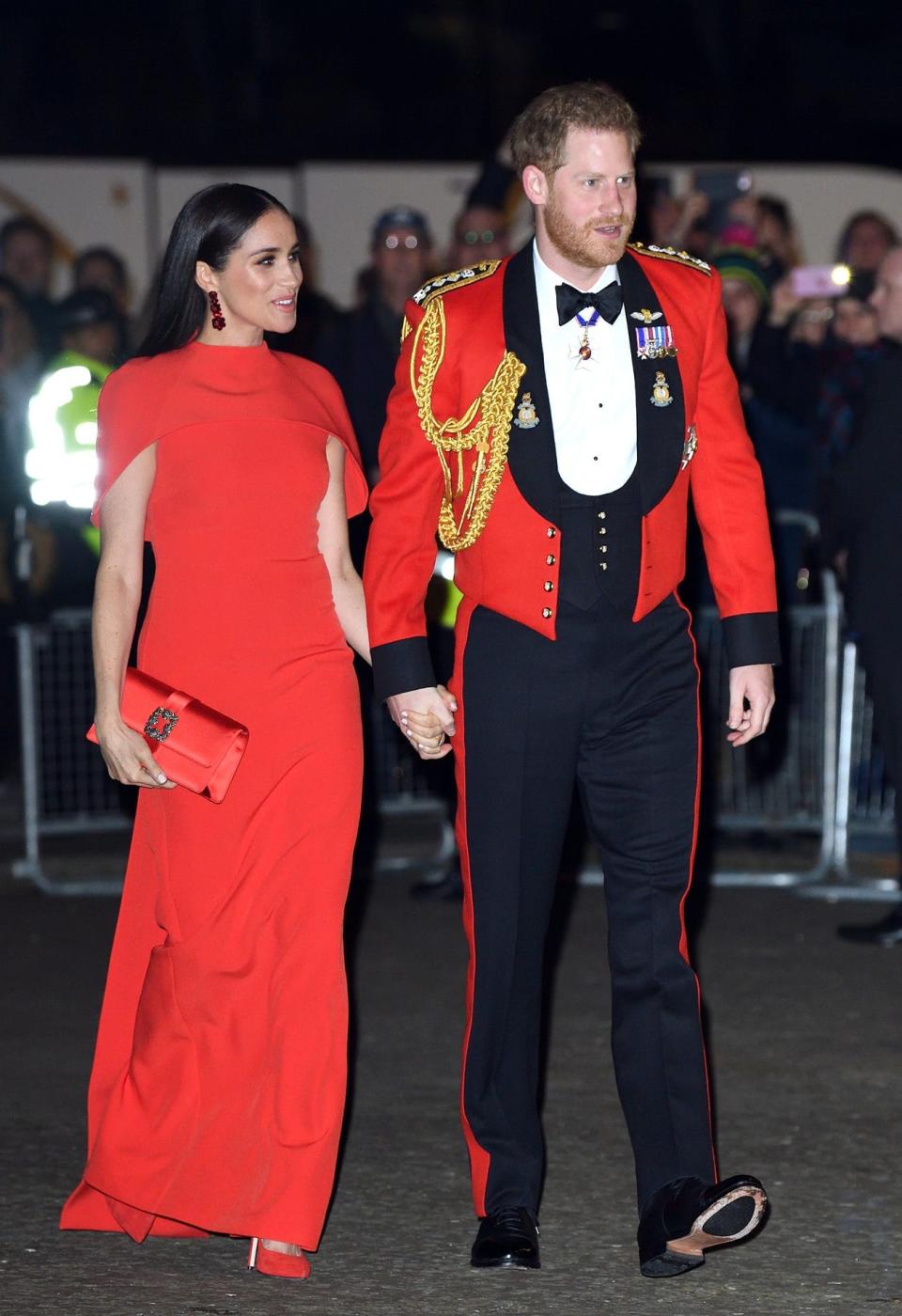 Meghan in a red cape sleeve dress and Harry in a red military jacket with badges, ropes, and a bow tie.