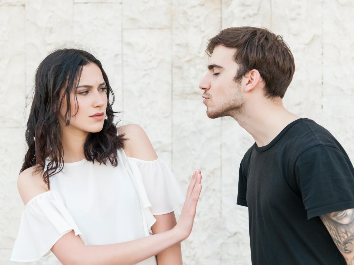 A young guy is leaning in to kiss a girl be she is frowning and making a hand gesture to stop him. They are wearing casual clothes.