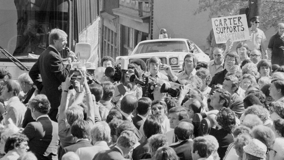 jimmy carter stands among a crowd and speaks into a microphone, behind him is a bus and police car