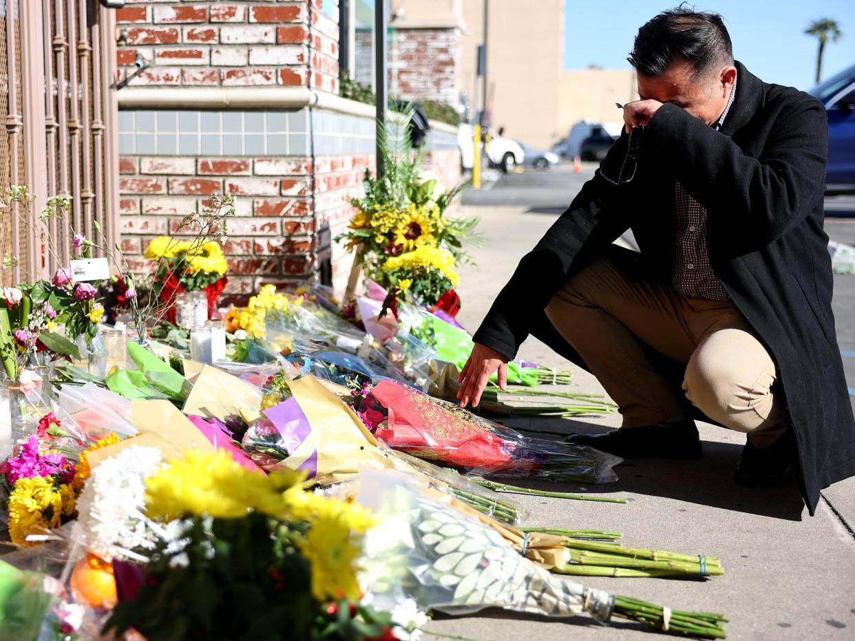 A man in a black coat and tan pants kneels in front of a gate with dozens of yellow flowers in front