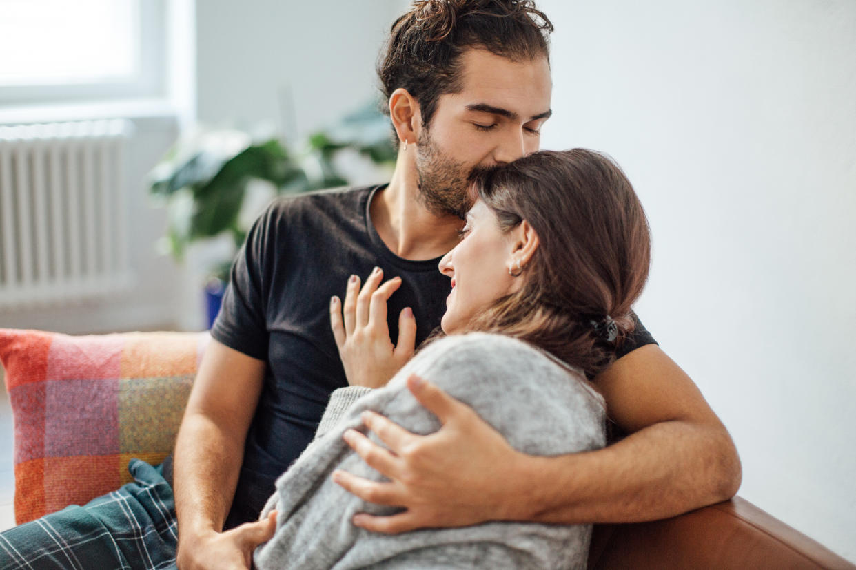 Young man embracing girlfriend while kissing on her forehead in living room at home love language