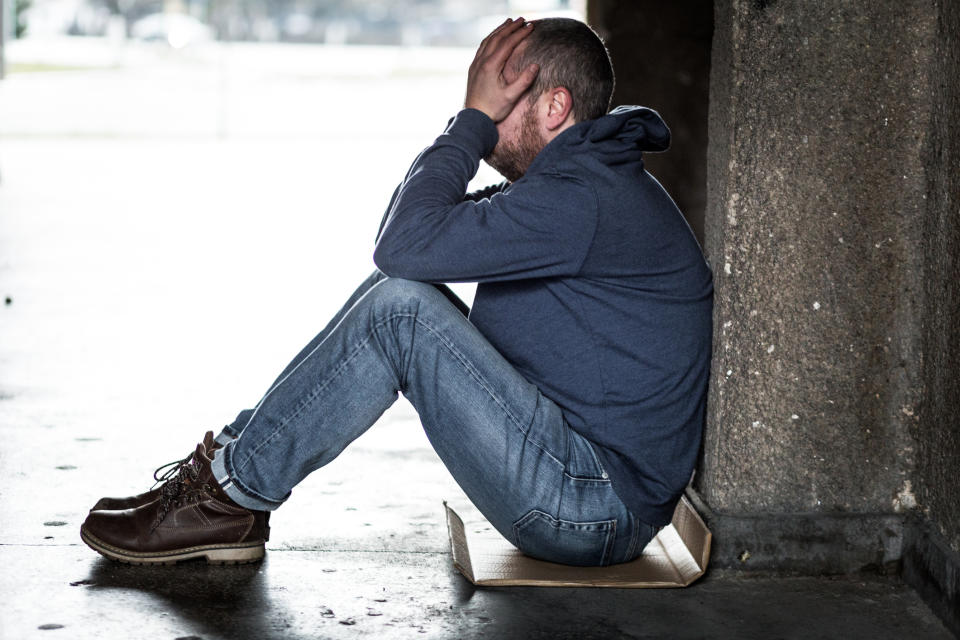 A man in a hoodie, jeans, and boots sits against a wall with his head in his hands, appearing distressed