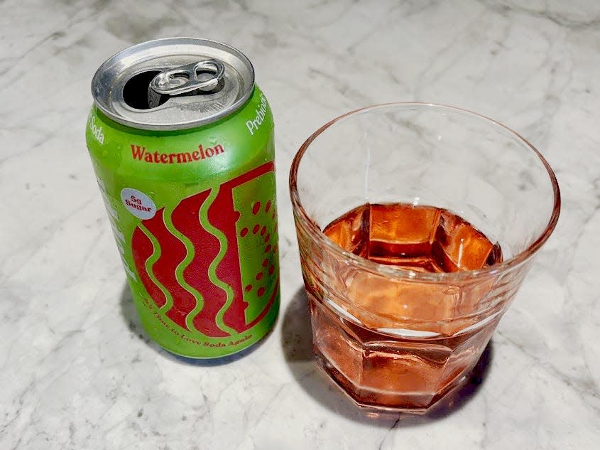 An open can of watermelon Poppi next to a small, clear glass with pink liquid inside. Both are sitting on a countertop.