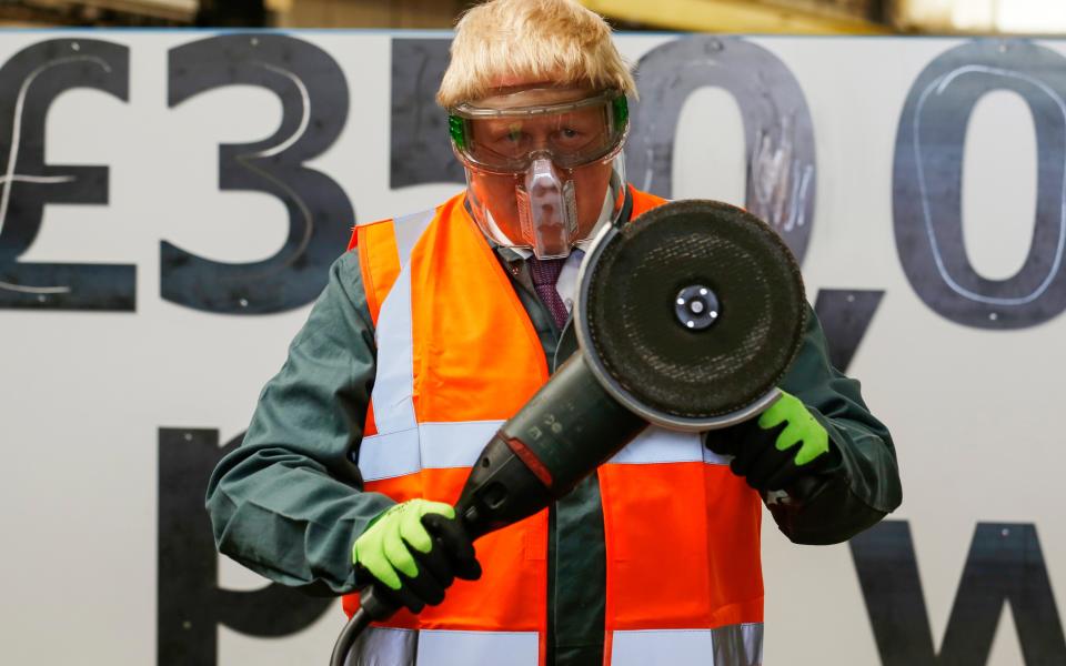 Boris Johnson, in a high-vis gear and a protective mask holds up a large angle grinder - Credit: Luke MacGregor/Bloomberg