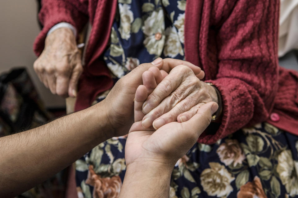 A younger person is holding the hands of an older person who is wearing a floral dress and a textured cardigan