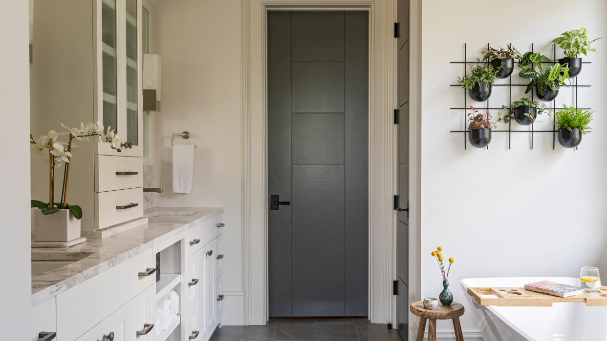  White bathroom with built-in cupboards, grey internal doors and plant grid on wall above white bath. 