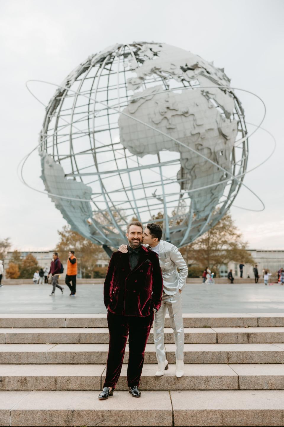 A groom kisses his husband on the cheek in front of a giant statue of a globe.