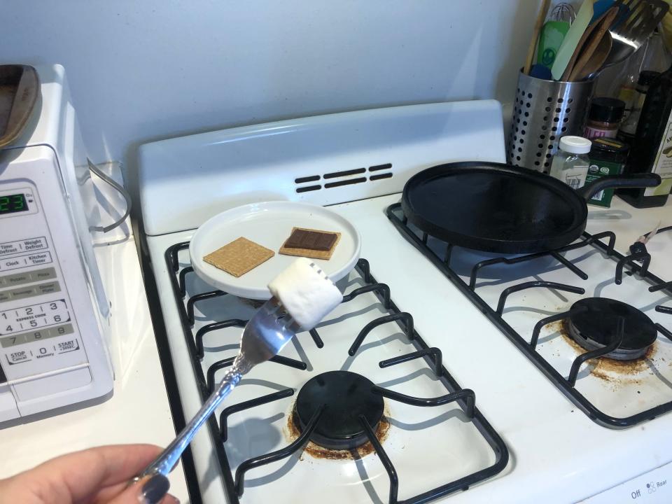 hand holding a fork with a marshmallow on it over a stove with a plate of graham crackers and chocolate in the background