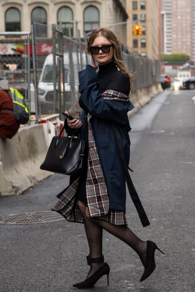 anna delvey looks at the camera while crossing the street, she wears a long navy and plaid jacket, a black turtleneck dress, black hose, black heels, and large black sunglasses, she holds her phone in one hand and has her purse on her wrist