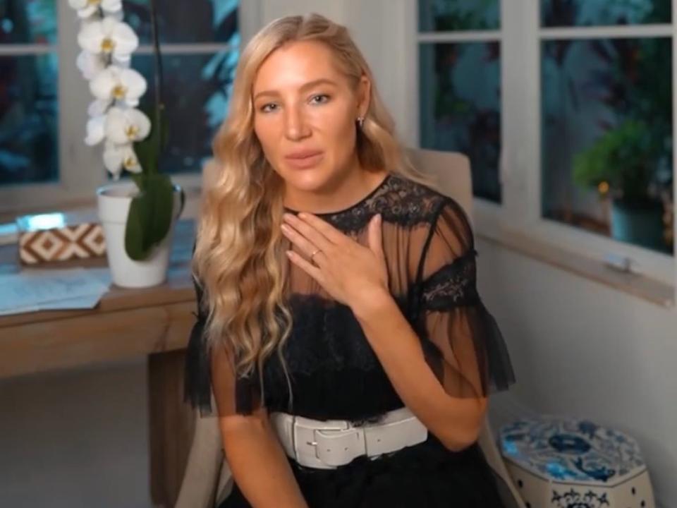 A woman sits in a room with windows and holds a hand to her chest.