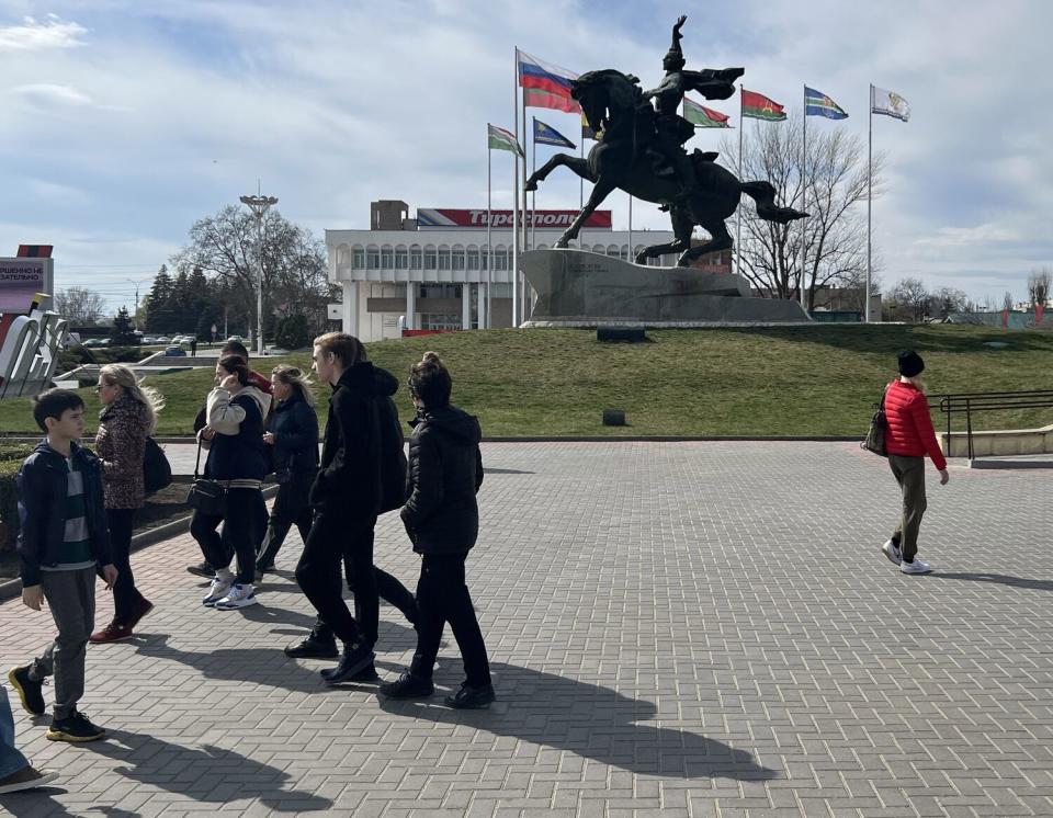 Families strolling a park with a statue of a person on horseback in the background.