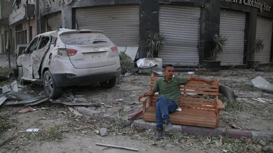 A man with medium-toned skin sits casually on a bench amid a scene of destruction with shuttered businesses and a car with crumpled doors, debris across the ground.