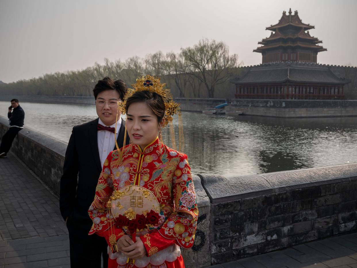 A bride dressed in a traditional Chinese wedding dress and a groom pose for pictures with the corner tower of the Forbidden City in the background in Beijing, China.