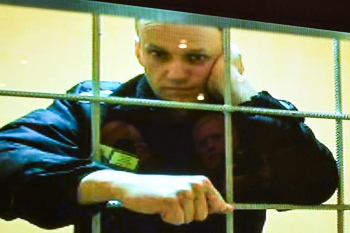 An image of Alexey Navalny, standing behind prison bars, appears on a screen in a courtroom.