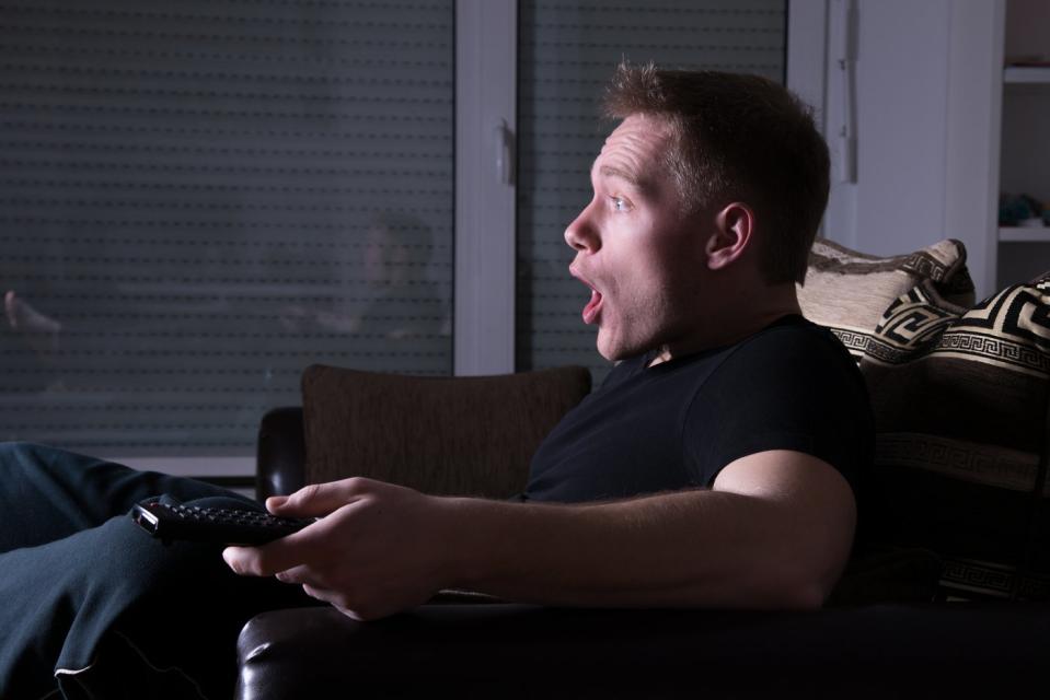 Individual on a sofa with a shocked expression, remote in hand, in a dimly lit living room.