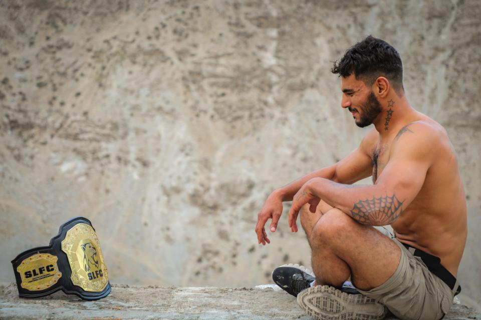 A man sits cross legged facing an gold MMA championship belt.