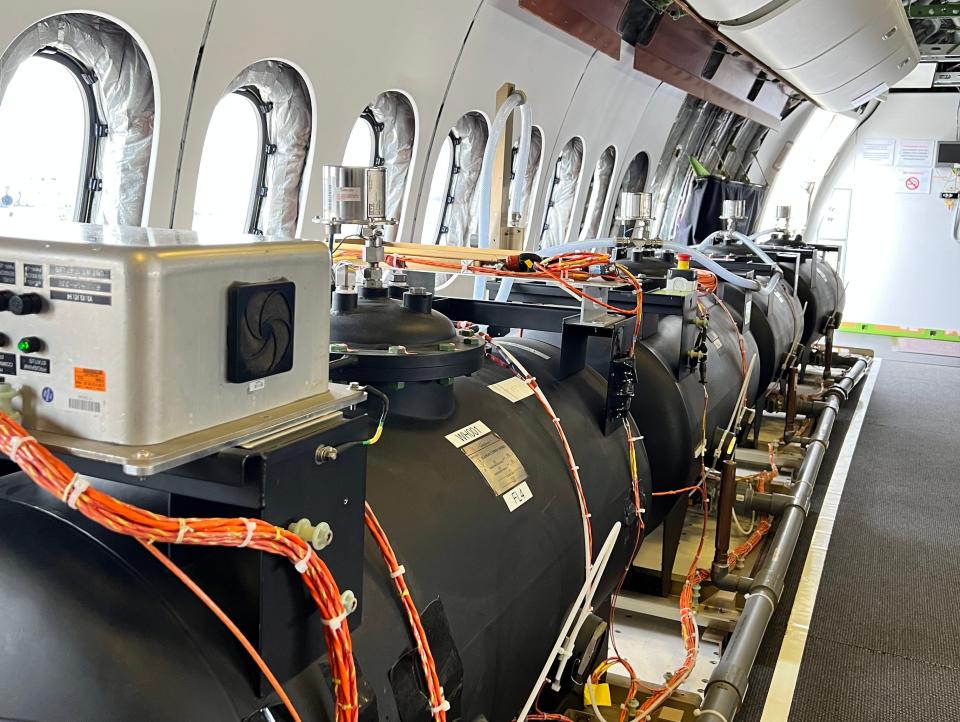 A row of black water tanks acting as ballasts onboard a Boeing 777-9 testbed, windows in the background and stripped-down walls