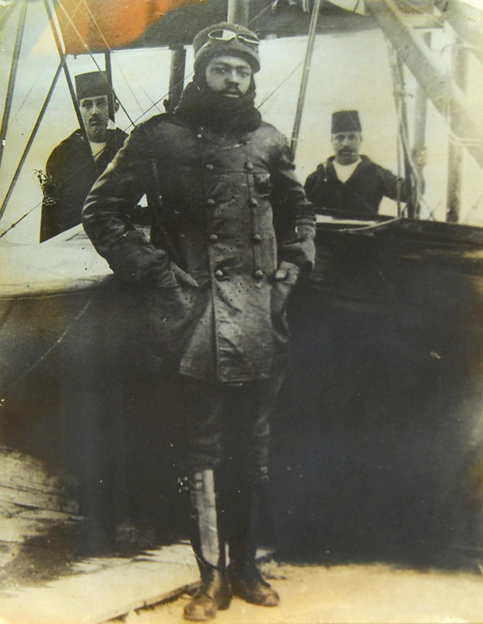 A man with high boots and in a uniform and aviator glasses stands in front of an early plane