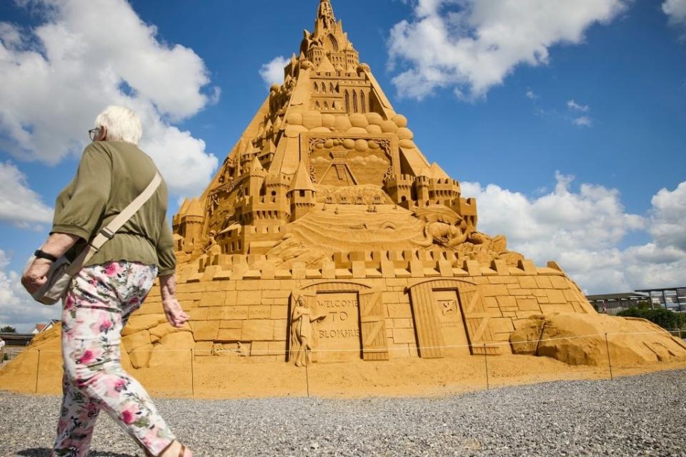 An elaborate sandcastle sculpture with intricate details, including doors, windows, and battlements, and a person walking by in the foreground