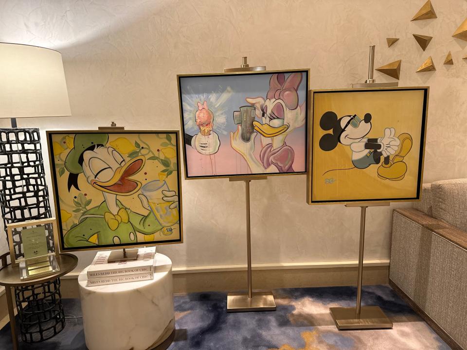 Three pieces of Disney art held up by gold frames on metal stands. The photo on the right shows Mickey with glasses sitting with his hands clasped around his knees against a yellow background. The painting in the middle is of Daisy Duck wearing a pink bow and a pink dress, taking a photo of a gloved hand holding a melting ice cream with a pink bow on top. The background is blue, fading to pink horizontally. The painting on the left, which rests on a pile of books on top of a round marble table, shows Donald Duck laughing while holding a glass of lemonade with ice. His eyes are closed, and he wears a light green suit and hat. He's surrounded by branches of a lemon tree against a yellow background.