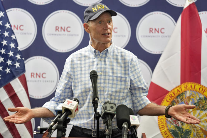 Sen. Rick Scott at the microphone, wearing a navy baseball cap printed with the word Navy, and RNC (Republican National Committee) seals in the background.