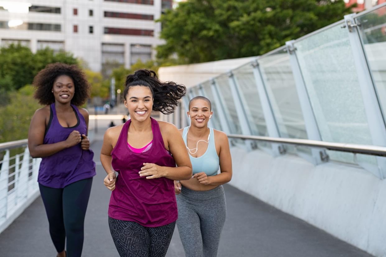 Happy young curvy women jogging together on city bridge. Healthy girls friends running on the city street to lose weight. Group of multiethnic oversize women running with building in the background.